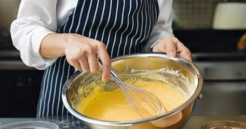 A professional chef gently folding Madeleine cookie batter with a spatula in a pristine kitchen